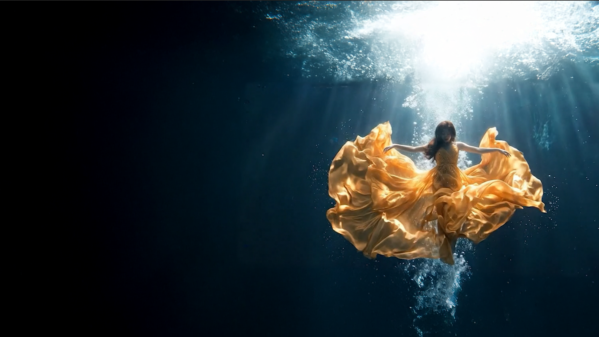 A woman in a golden dress suspended underwater, light streaming down